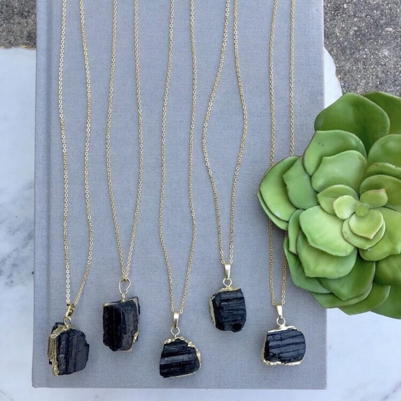 May include: Five gold-coloured necklaces featuring raw black tourmaline pendants. The pendants have gold-coloured edges and hang from delicate gold chains. The necklaces are displayed on a grey surface next to a green succulent.