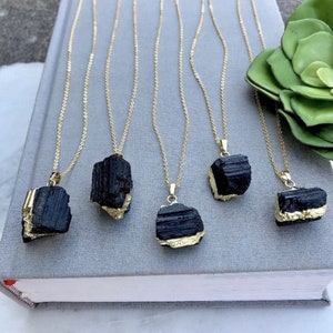 May include: Five gold-coloured necklaces featuring black tourmaline pendants. Each pendant has a raw, natural shape with gold-coloured accents. The necklaces are displayed on a grey surface, with a green plant visible in the background.