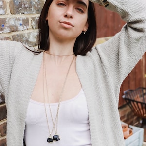 May include: A woman wearing a light grey cardigan over a white ribbed vest top. She is wearing two gold necklaces, each with a black pendant. A gold bracelet is on her left wrist. The background includes a brick wall and greenery.
