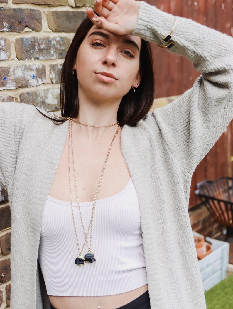 May include: A woman wearing a light grey cardigan over a white ribbed vest top. She is wearing two gold necklaces, each with a black pendant. A gold bracelet is on her left wrist. The background includes a brick wall and greenery.