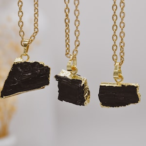 May include: Three gold-coloured chain necklaces, each with a black, irregularly shaped pendant. The pendants have gold-coloured edges. The necklaces are displayed against a soft, neutral background.