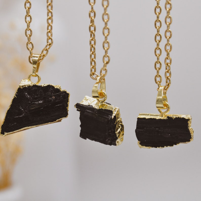 May include: Three gold-coloured chain necklaces, each with a black, irregularly shaped pendant. The pendants have gold-coloured edges. The necklaces are displayed against a soft, neutral background.