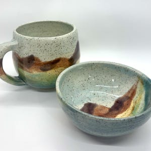 May include: A ceramic mug and a matching bowl featuring a speckled off-white glaze with horizontal bands of brown, yellow, and teal. The mug has a curved handle, and both pieces exhibit a hand-thrown appearance with a smooth, glossy finish.