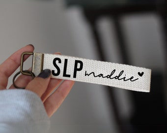 May include: White canvas keychain with black text that reads "SLP maddie" with a small black heart.