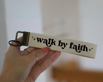 Peut inclure: Un porte-clés en toile blanche avec un anneau en métal brun et le texte "walk by faith" en noir.