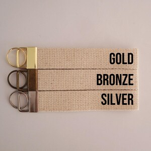 Op de afbeelding: Drie beige canvas sleutelhangers met metalen hardware in goud, brons en zilver. De tekst "GOLD", "BRONZE" en "SILVER" is in zwart op het canvas gedrukt.