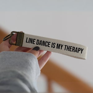 Puede incluir: Un llavero de lona blanco con un anillo de metal marrón y el texto "LINE DANCE IS MY THERAPY" impreso en negro.