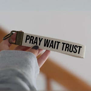 Op de afbeelding: Een witte canvas sleutelhanger met een messing sleutelring en de tekst "Pray Wait Trust" in zwarte letters.