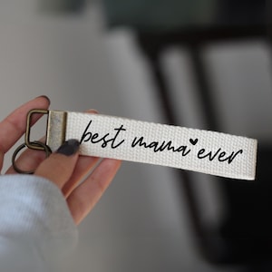May include: White canvas keychain with a black inscription that reads "best mama ever" with a small heart symbol.