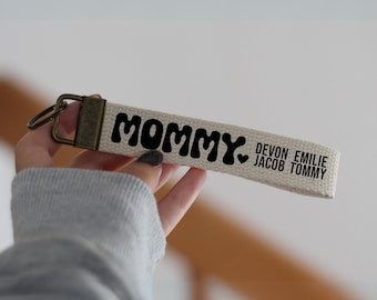 Op de afbeelding: Witte canvas sleutelhanger met het woord "Mommy" in zwarte, golvende letters. De sleutelhanger heeft ook de namen "Devon, Emilie, Jacob, Tommy" in zwart.