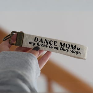 Op de afbeelding: Witte canvas sleutelhanger met zwarte tekst die "Dance Mom my heart is on that stage" luidt met een klein zwart hart symbool.