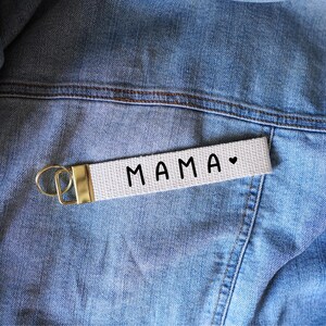 Op de afbeelding: Een witte canvas sleutelhanger met het woord "MAMA" in zwarte letters en een klein hartsymbool. De sleutelhanger heeft een goudkleurige metalen sluiting en wordt getoond op een spijkerjasje.