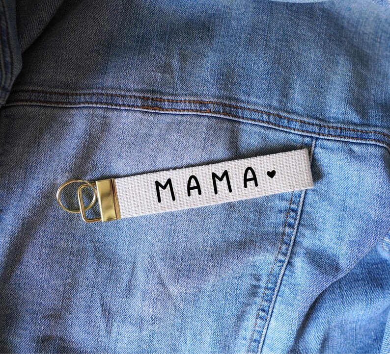 Op de afbeelding: Een witte canvas sleutelhanger met het woord "MAMA" in zwarte letters en een klein hartsymbool. De sleutelhanger heeft een goudkleurige metalen sluiting en wordt getoond op een spijkerjasje.