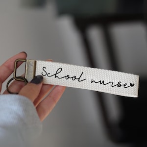 Könnte beinhalten: Ein cremefarbener Schlüsselanhänger mit dem Schriftzug "School nurses" in schwarzer Schrift. Der Schlüsselanhänger hat einen bronzefarbenen Metallverschluss und -ring. Der Stoff ist ein gewebtes Material.