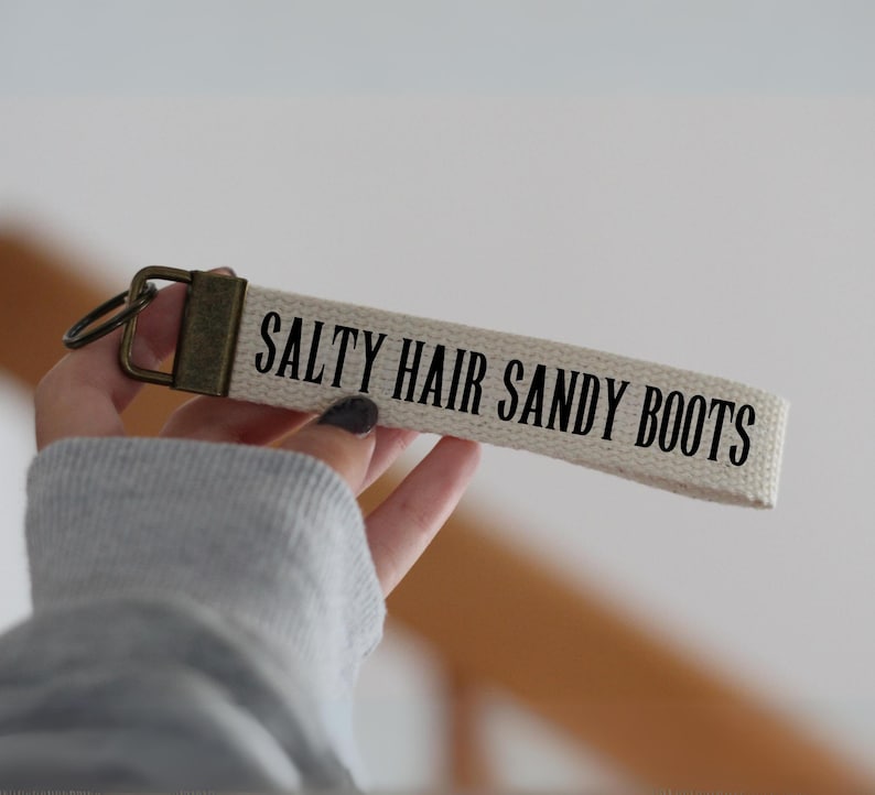 May include: A beige canvas keychain wristlet with the text "SALTY HAIR SANDY BOOTS" in black. The keychain has a bronze-colored metal clasp and ring.