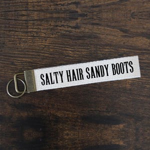 May include: A white canvas keychain strap with the text "SALTY HAIR SANDY BOOTS" in black lettering. The strap has a metal clasp and key ring. The background is a dark brown wooden surface.
