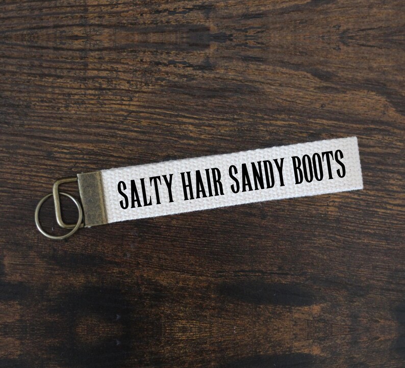 May include: A white canvas keychain strap with the text "SALTY HAIR SANDY BOOTS" in black lettering. The strap has a metal clasp and key ring. The background is a dark brown wooden surface.