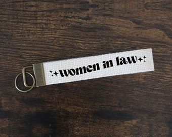 May include: A white canvas keychain with the text "women in law" in black, accented with starburst graphics. The keychain has a bronze-colored metal clasp and key ring, set against a dark wood grain background.