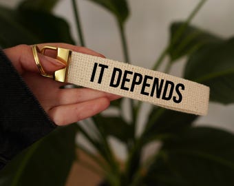 May include: A beige canvas keychain with a gold metal ring and the text "IT DEPENDS" printed in black.