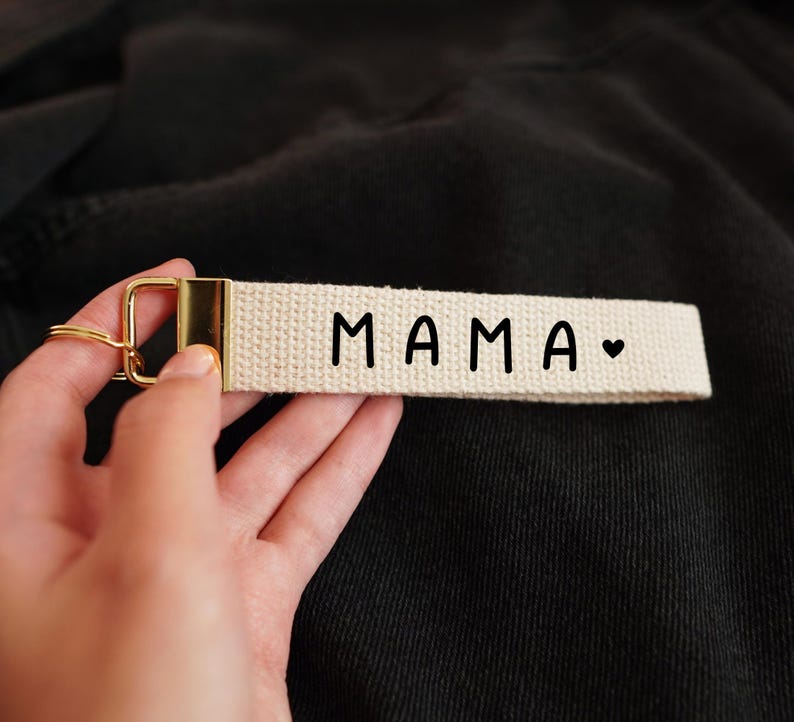 Op de afbeelding: Een cr&egrave;mekleurige sleutelhanger met het woord "MAMA" in zwarte letters en een klein hartsymbool. De band heeft een goudkleurige metalen sluiting en sleutelring.