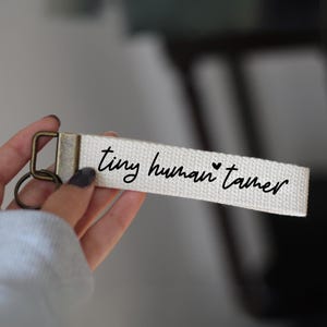 以下が含まれることがあります： 小さなハートのシンボルが付いた「tiny human tamer」という黒い文字が書かれた白いキャンバスのキーホルダーです。