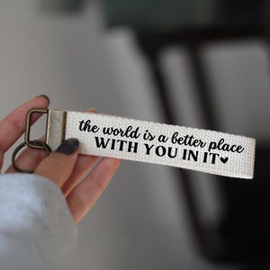 May include: White canvas keychain with a black inscription that reads "the world is a better place WITH YOU IN IT" with a small black heart symbol.