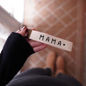 Op de afbeelding: Een cr&egrave;mekleurige sleutelhanger met het woord "MAMA" in zwarte letters, gevolgd door een klein hartje. De sleutelhanger heeft een bronzen metalen ring en een bijpassende bronzen metalen sluiting.