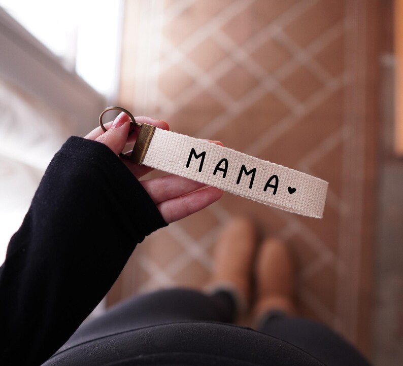 Op de afbeelding: Een cr&egrave;mekleurige sleutelhanger met het woord "MAMA" in zwarte letters, gevolgd door een klein hartje. De sleutelhanger heeft een bronzen metalen ring en een bijpassende bronzen metalen sluiting.