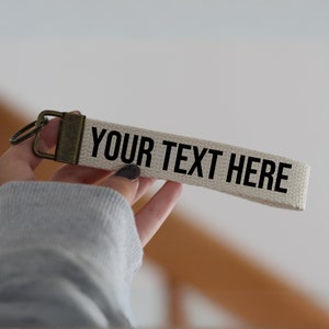 May include: A white canvas keychain with a brass key ring and the text "YOUR TEXT HERE" printed in black.