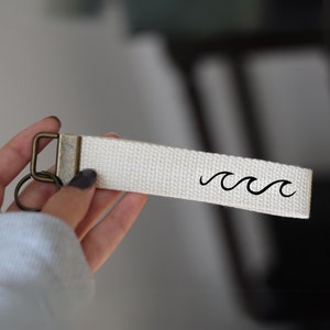 Könnte beinhalten: Weißer Canvas-Schlüsselanhänger mit einem Messing-Schlüsselring und einem schwarzen Wellendesign.