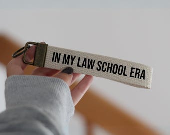 May include: A white canvas keychain with a brown metal ring and the text "IN MY LAW SCHOOL ERA" in black.