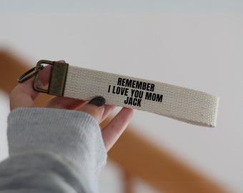 Op de afbeelding: Witte canvas sleutelhanger met een zwart geborduurd bericht dat "Remember I Love You Mom Jack" luidt.