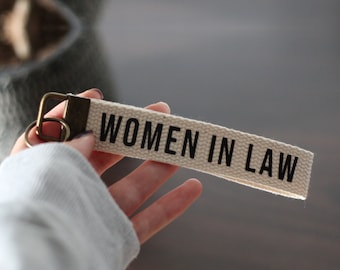 May include: A canvas keychain with the text "WOMEN IN LAW" printed in black. The keychain has a bronze-colored metal ring and clasp. The canvas is a natural, off-white color.