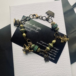 May include: A gold-toned charm bracelet with a teapot, crown, and teacup charms. The bracelet also features green and gold beads. The bracelet is on a white surface with a black business card that says "andco designs" and includes contact information.