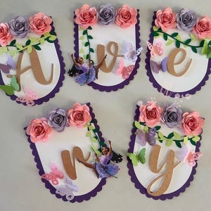 May include: A banner spelling out "AVERY" with paper flowers, butterflies, and a cartoon character. Each letter is on a white, scalloped-edged banner with a purple border. The flowers are pink and purple.