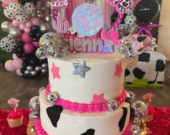 May include: A two-tiered white cake decorated with pink frosting, black and white cow print, silver disco balls, and pink stars. The cake is topped with a pink and white cake topper that says "One" and "Sienna".