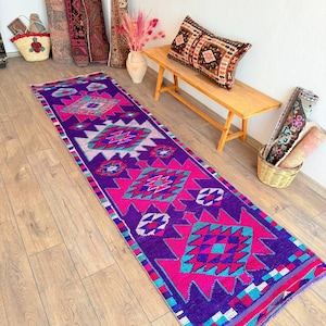 May include: A long, rectangular runner rug with a vibrant geometric pattern in shades of purple, pink, and turquoise. The rug is placed on a wooden floor, showcasing its intricate design and color palette. Other rugs and decorative items are in the background.
