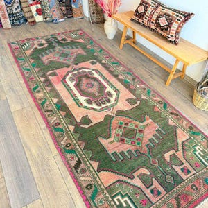 Op de afbeelding: Een rechthoekig vloerkleed met een centraal geometrisch medaillonpatroon in tinten bosgroen, zachtroze en crème. Het kleed heeft een decoratieve rand met ingewikkelde motieven en een gefranjerde rand, met een verweerde, versleten textuur.