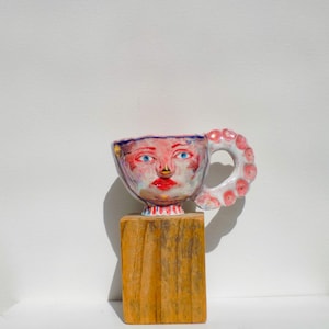 Puede incluir: Una taza de cerámica caprichosa con una cara pintada, con ojos azules, labios rojos y una nariz dorada. El asa está adornada con lunares rosas. La taza descansa sobre un pequeño bloque de madera rectangular, creando una pieza decorativa única.