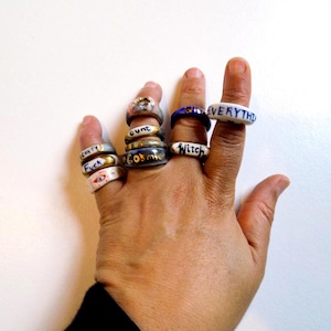May include: A hand wearing several rings with various designs and text. The rings are in different colors, including white, blue, gold, and silver. The text on the rings includes words like "Cosmic", "Witch", and "Everything".