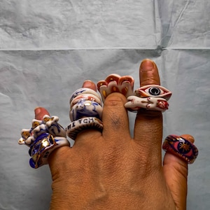 May include: A collection of ceramic rings worn on fingers. The rings feature various designs including a blue and white pattern with the text WITCH, a ring with a central eye motif, a ring with a gold star, and other colorful, sculpted shapes.