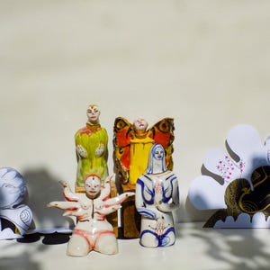 May include: Four ceramic figurines, each with a unique design. One figurine is a woman with multiple arms, another is a woman with a butterfly wing design, another is a woman with a yellow dress, and the last is a woman with a blue dress.