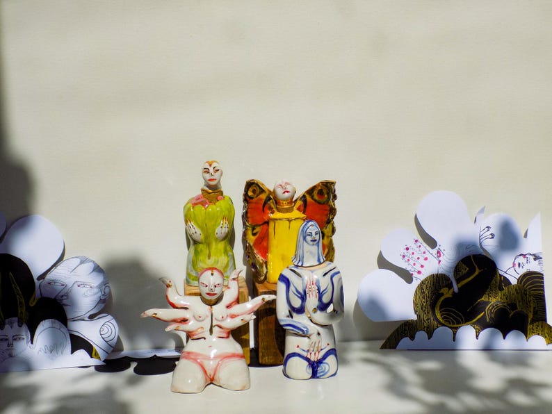 May include: Four ceramic figurines, each with a unique design. One figurine is a woman with multiple arms, another is a woman with a butterfly wing design, another is a woman with a yellow dress, and the last is a woman with a blue dress.
