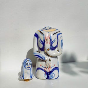 May include: A white ceramic figurine of a person kneeling with blue and gold painted details. The figurine has a stylized face and body. A smaller white ceramic figurine of a person's head with blue hair is also shown.