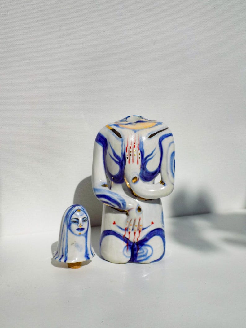 May include: A white ceramic figurine of a person kneeling with blue and gold painted details. The figurine has a stylized face and body. A smaller white ceramic figurine of a person's head with blue hair is also shown.