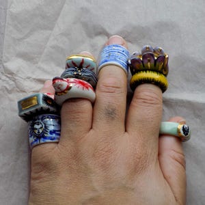 May include: A hand wearing several ceramic rings. The rings feature various designs, including a sun, a lotus flower, and geometric patterns. Colors include blue, yellow, red, and gold. The rings are of varying widths and styles.