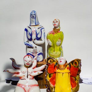 May include: Four ceramic figurines, each with a unique design. One figurine is a woman with blue and white painted clothing, another is a woman with green and yellow painted clothing, another is a woman with a bird and multiple arms, and the last is a woman with butterfly wings and yellow painted clothing.