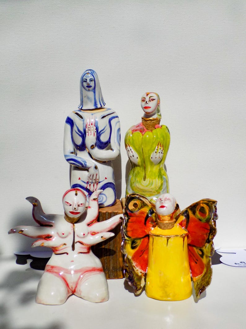 May include: Four ceramic figurines, each with a unique design. One figurine is a woman with blue and white painted clothing, another is a woman with green and yellow painted clothing, another is a woman with a bird and multiple arms, and the last is a woman with butterfly wings and yellow painted clothing.
