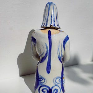 May include: A ceramic figurine of a person wearing a blue and white patterned dress and a blue and white striped hat. The figurine is standing on a white surface.