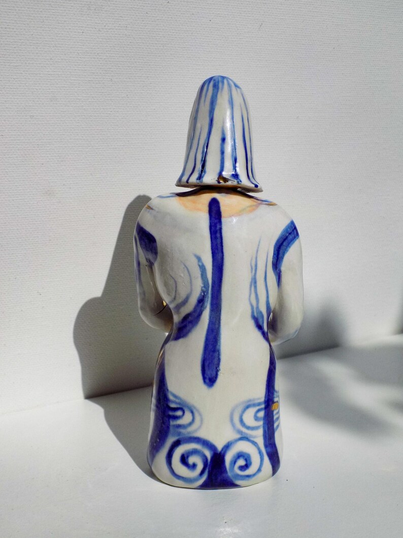 May include: A ceramic figurine of a person wearing a blue and white patterned dress and a blue and white striped hat. The figurine is standing on a white surface.